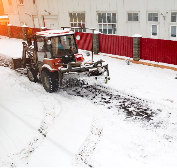 Tractor with snow sweeping attachment clearing snow-covered streets near commercial buildings.