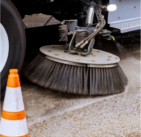 Close-up of a street sweeper brush cleaning pavement and debris near a traffic cone.